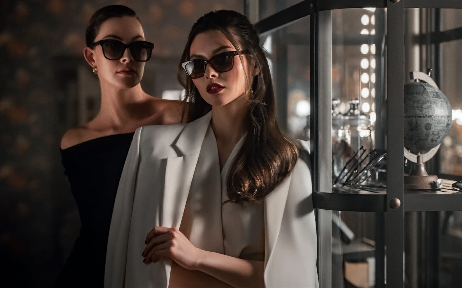 Two elegant women are standing next to a glass showcase. One is wearing a black strapless dress and the other a white suite. Both are wearing dark sunglasses.