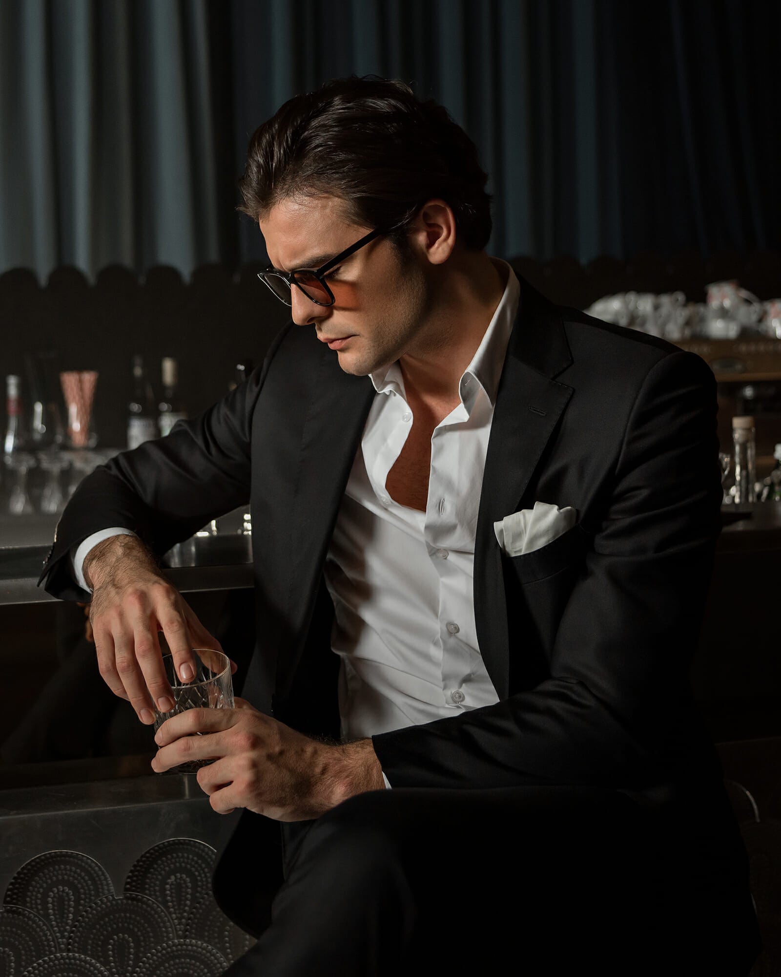 A dark haired man at an elegant bar. He is holding a whisky glass with his two hands, lowered gaze. He is wearing a black suit, a white shirt and black acetat sunglasses.