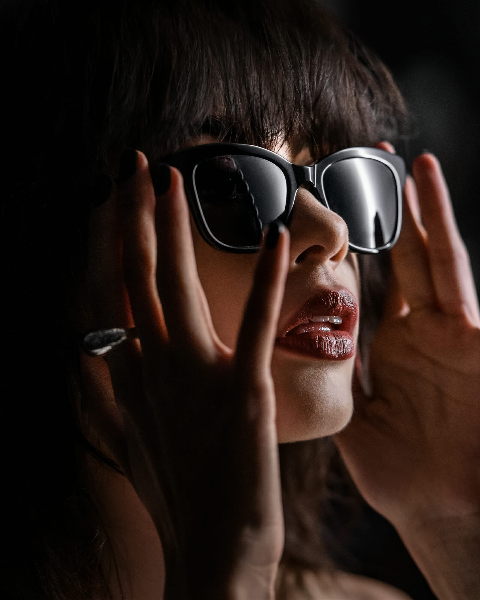 Close up of a dark haired woman who is looking to the side and holding her black acetat sunglasses. She is wearing a golden fingerring and red lipstick.