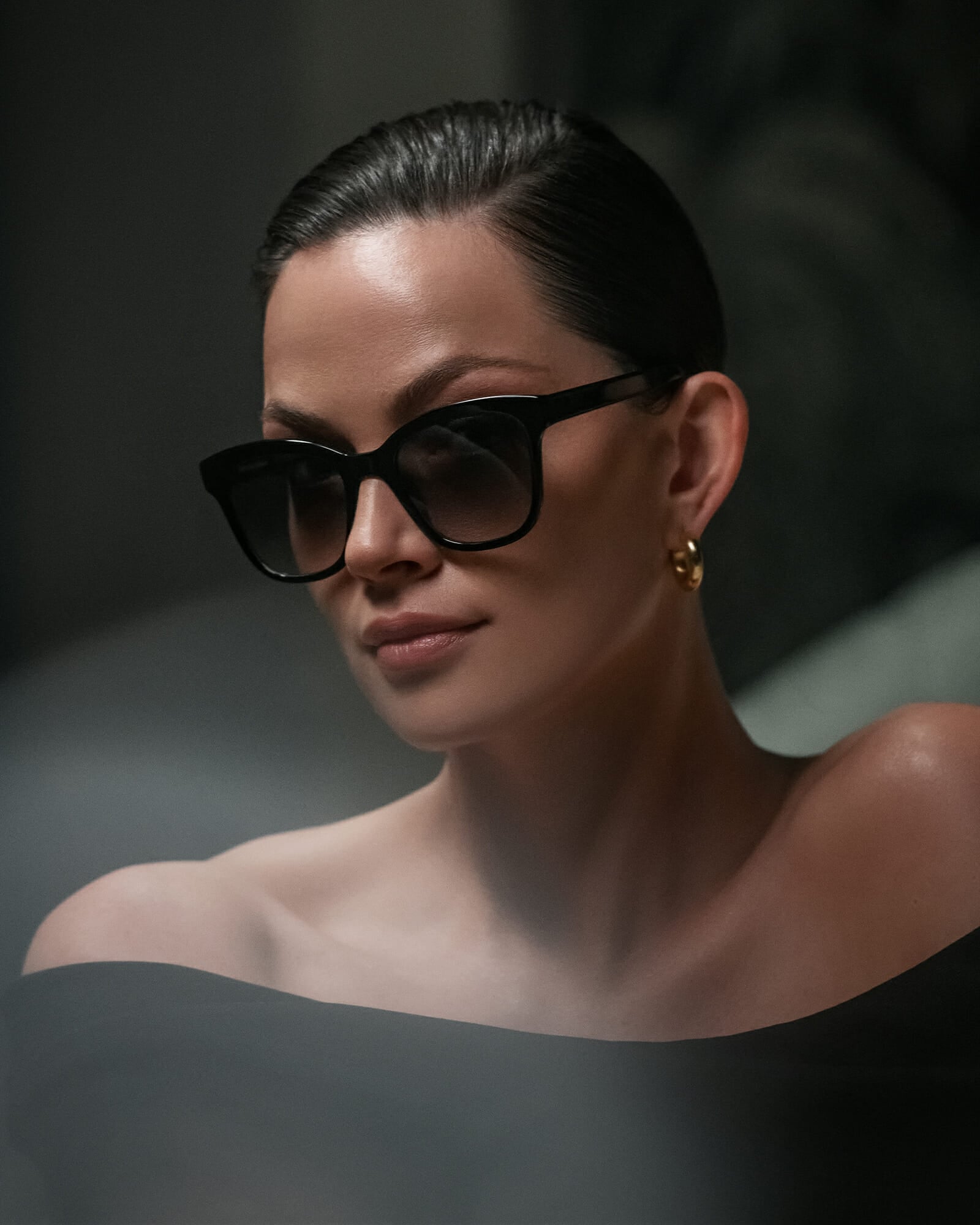 Portrait of an elegant brown haired woman who is looking into the camera. She is wearing black acetat sunglasses and e black strapless dress and golden earrings.