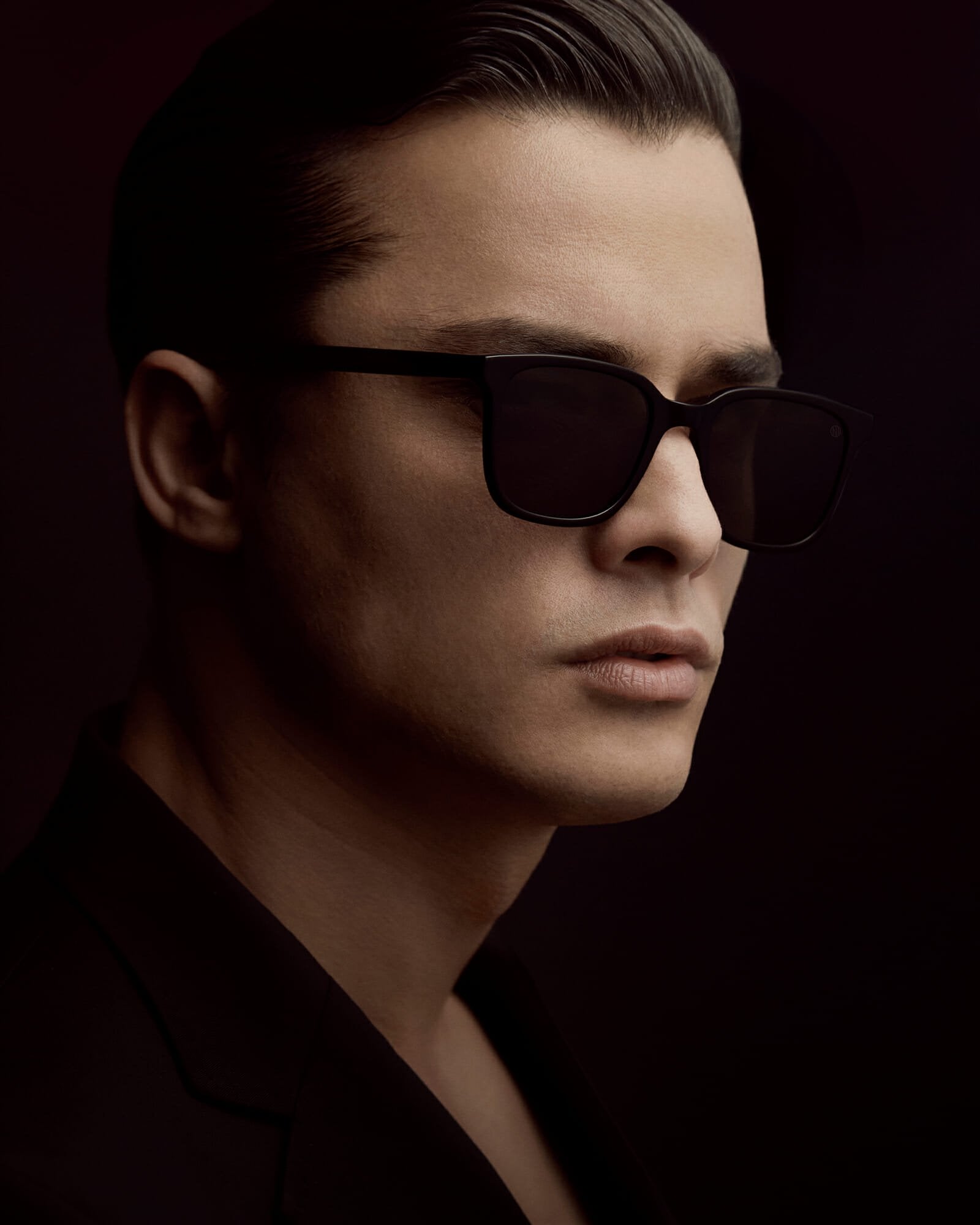 Portrait of a dark haired man who is wearing a black suit and black sunglasses. Viewed from the side.