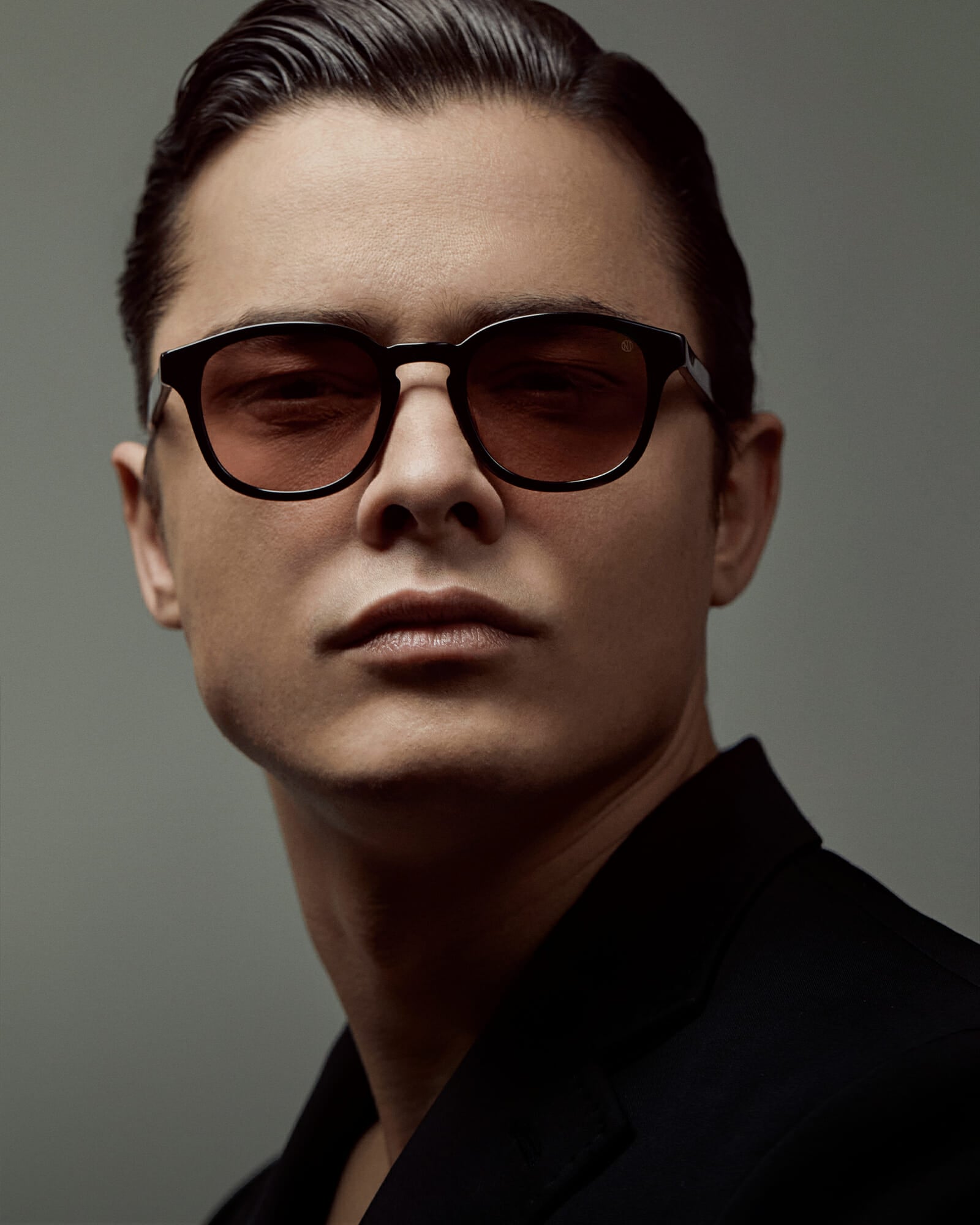 Portrait of a dark haired man who is wearing a black suit and black sunglasses. He is looking into the camera.